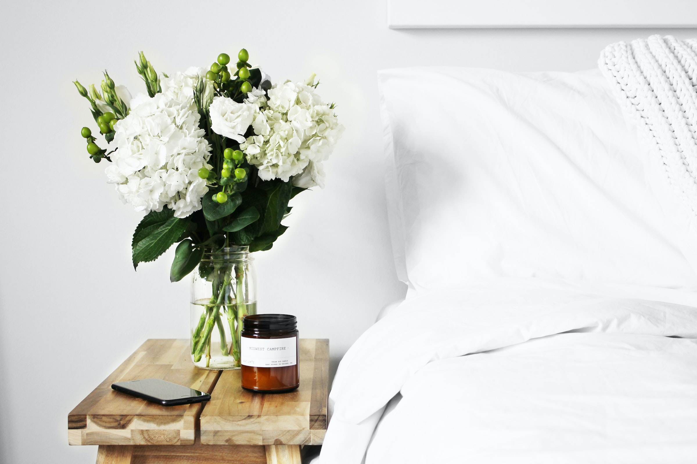 Flowers in a vase with candle and iPhone on a bedside table next to a bed