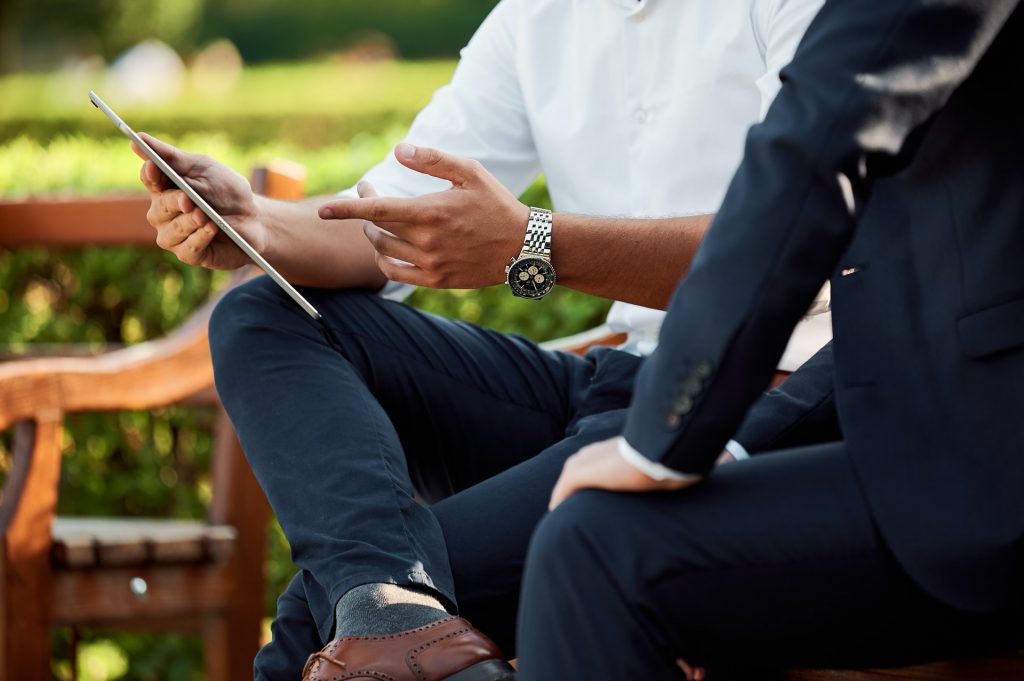 two men looking at an iPad, one man wearing a white shirt, wearing a stainless steel watch, the other man is wearing a dark suit