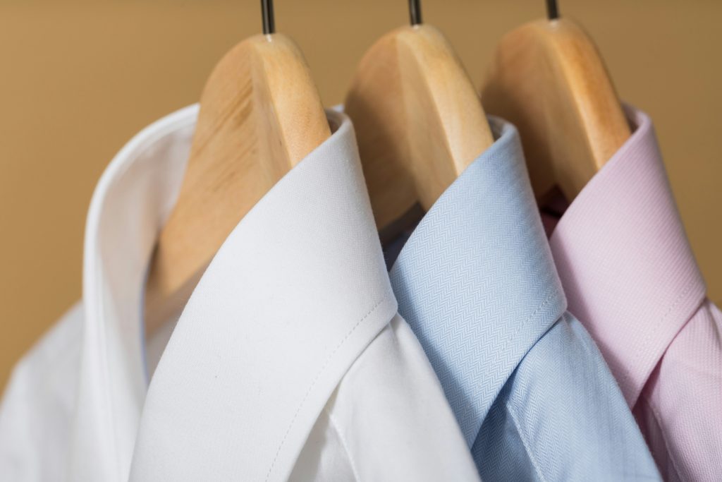 3 mens shirts hanging on wooden hangers. One of each; one in white, one in blue and pink.