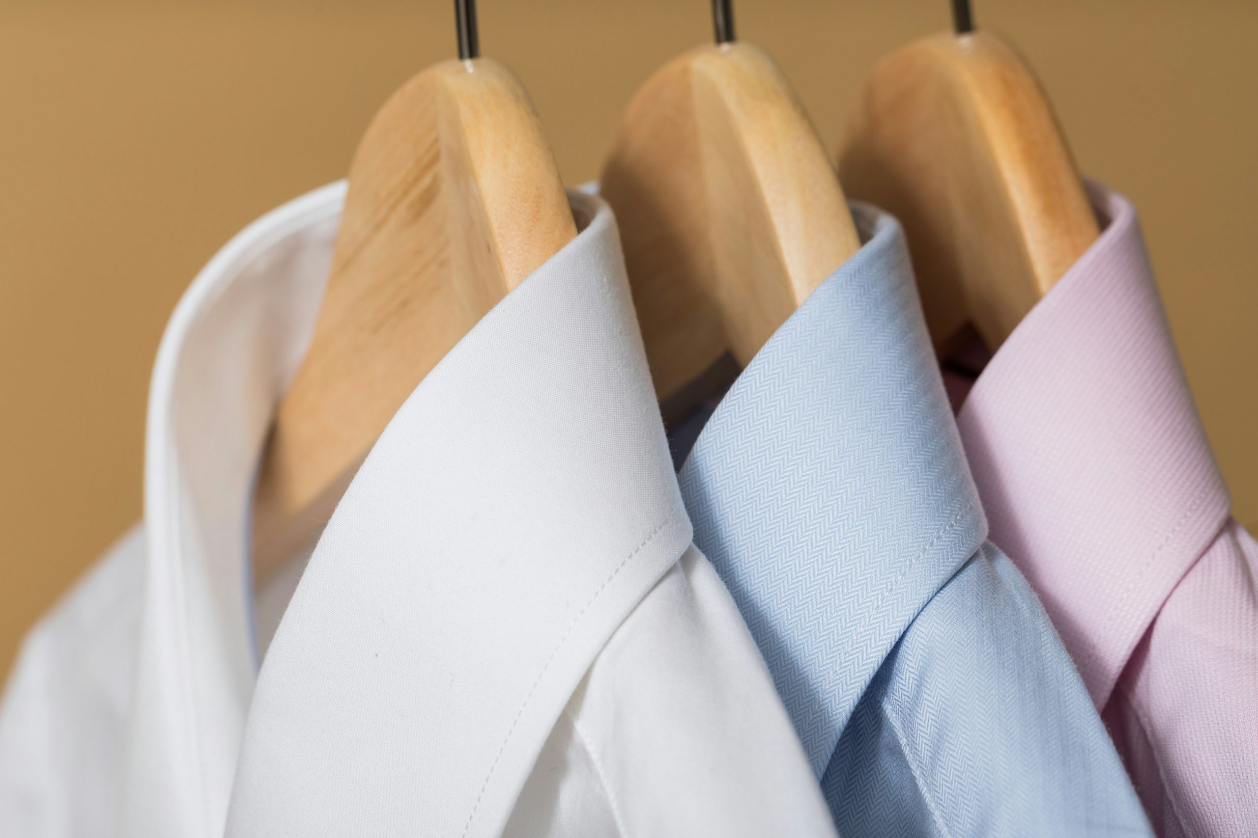 3 mens shirts hanging on wooden hangers. One of each; one in white, one in blue and pink.