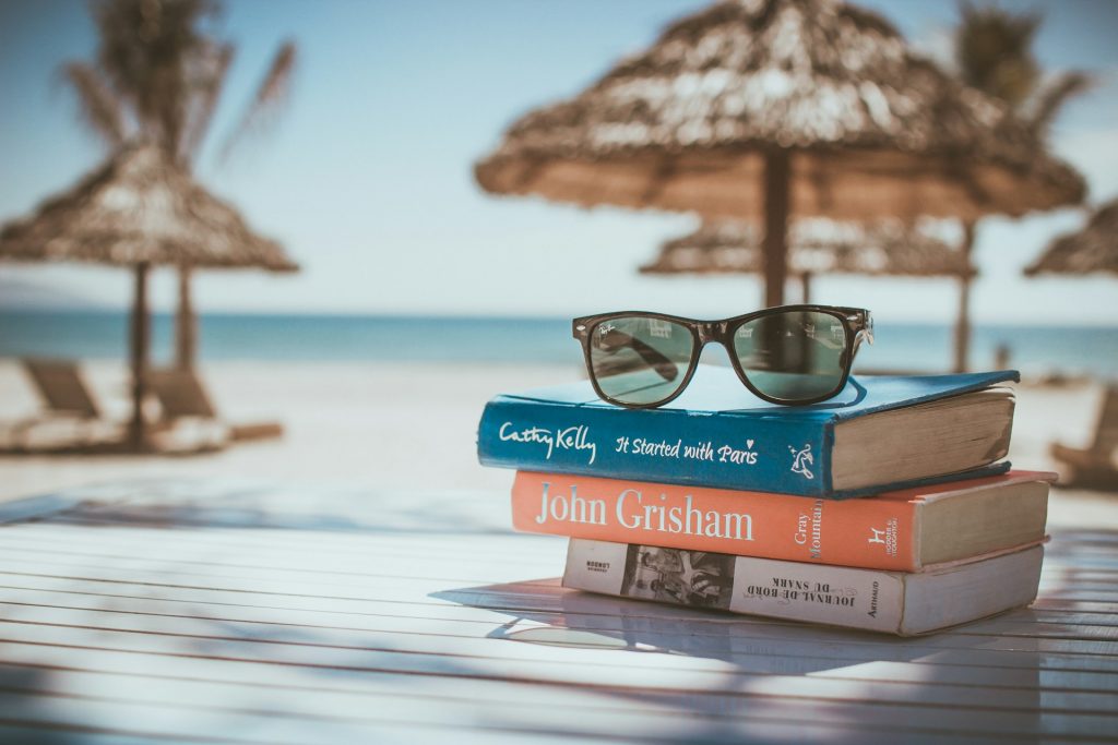 stack of books on the beach with sunglasses on top and an umbrella in the background