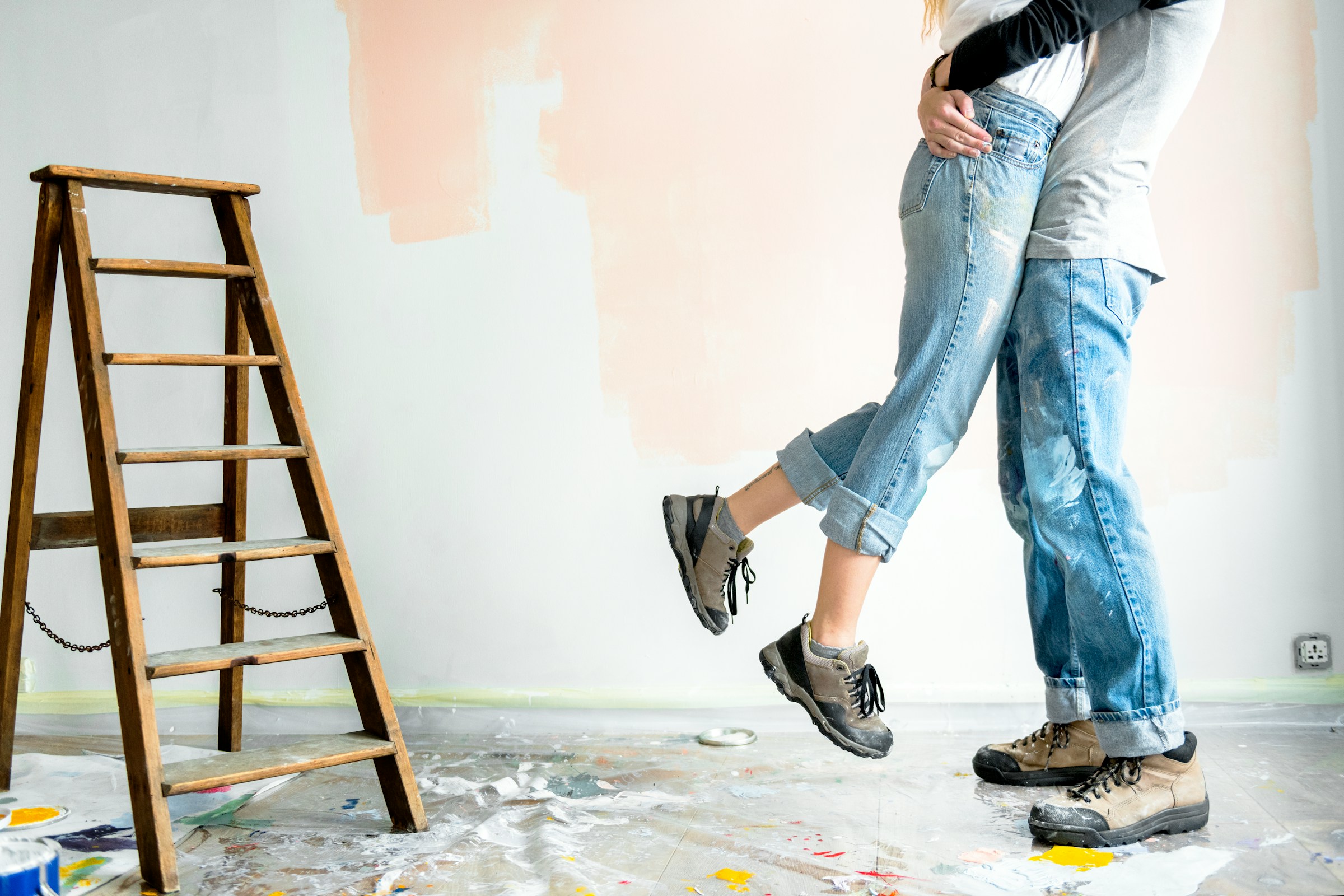 Man picking up a woman. Both are wearing paint covered pants with a ladder in the background.